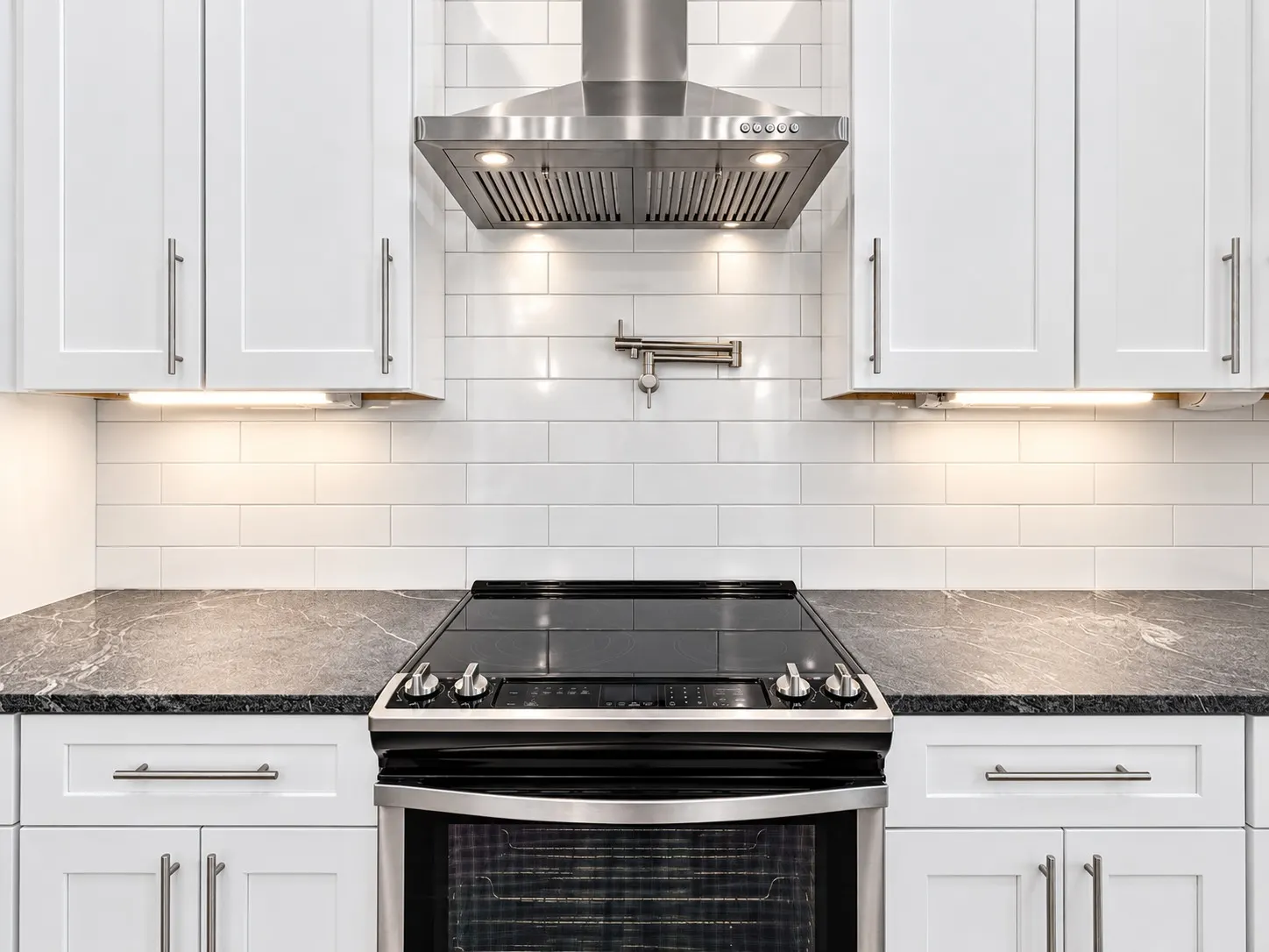 Kitchen range and stainless steel hood with white subway tile, a pot filler and black stone counters.
