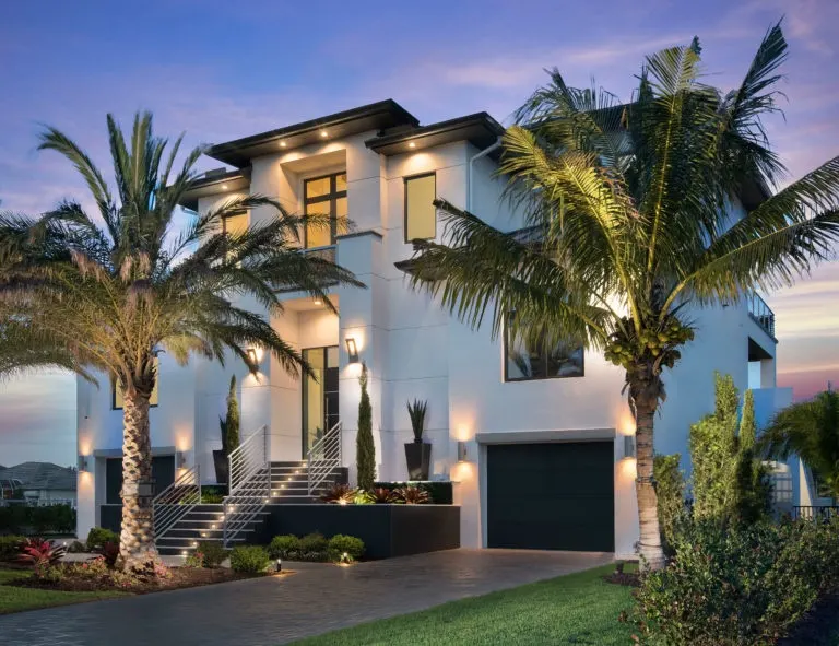 Front of the Vanderbilt Coastal Residence at dusk, a multi-level white contemporary home with an illuminated entry stair, palms and a paver driveway.
