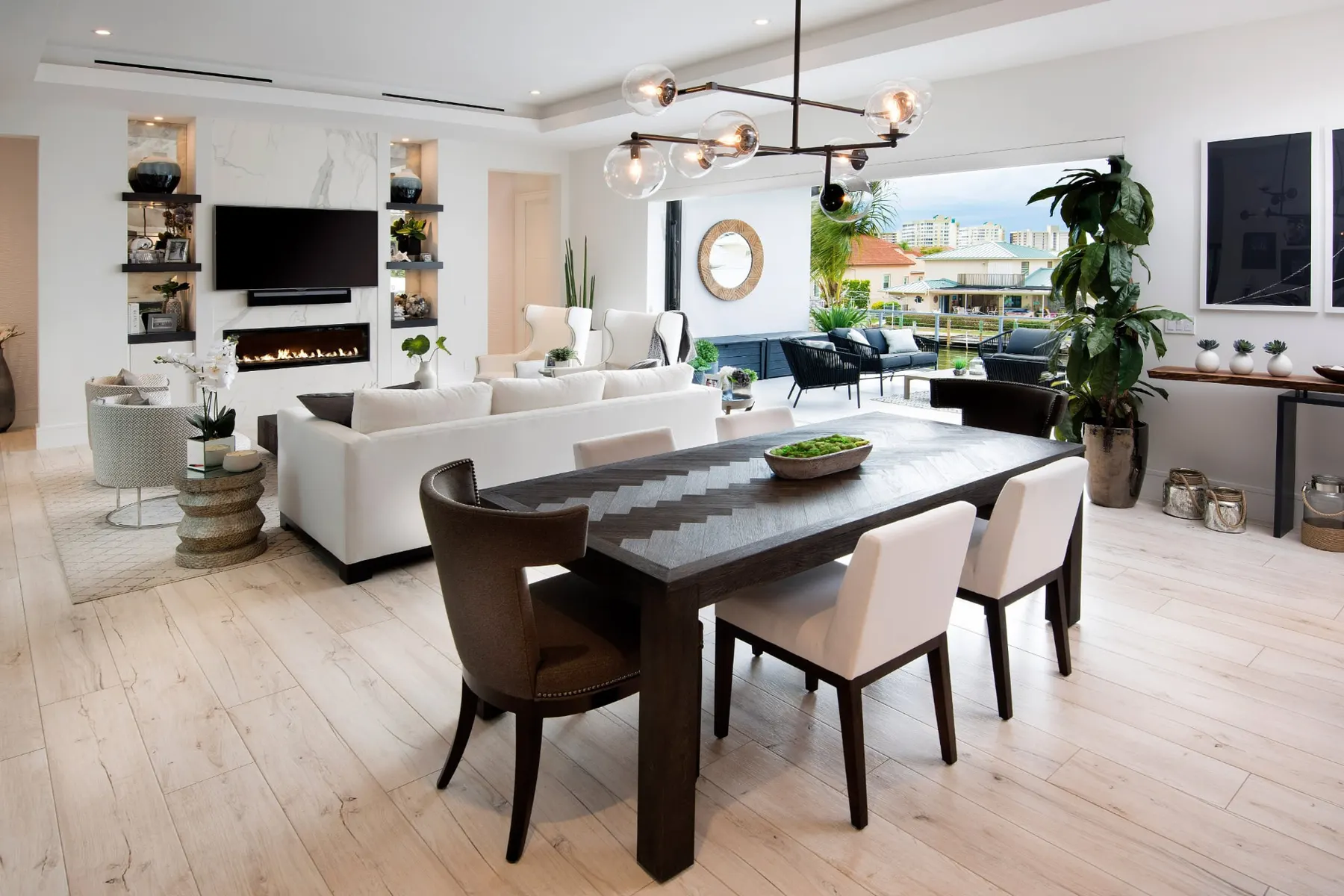 Open living and dining area with a linear fireplace in a marble-look wall, a dark herringbone dining table and a wide opening to the terrace.