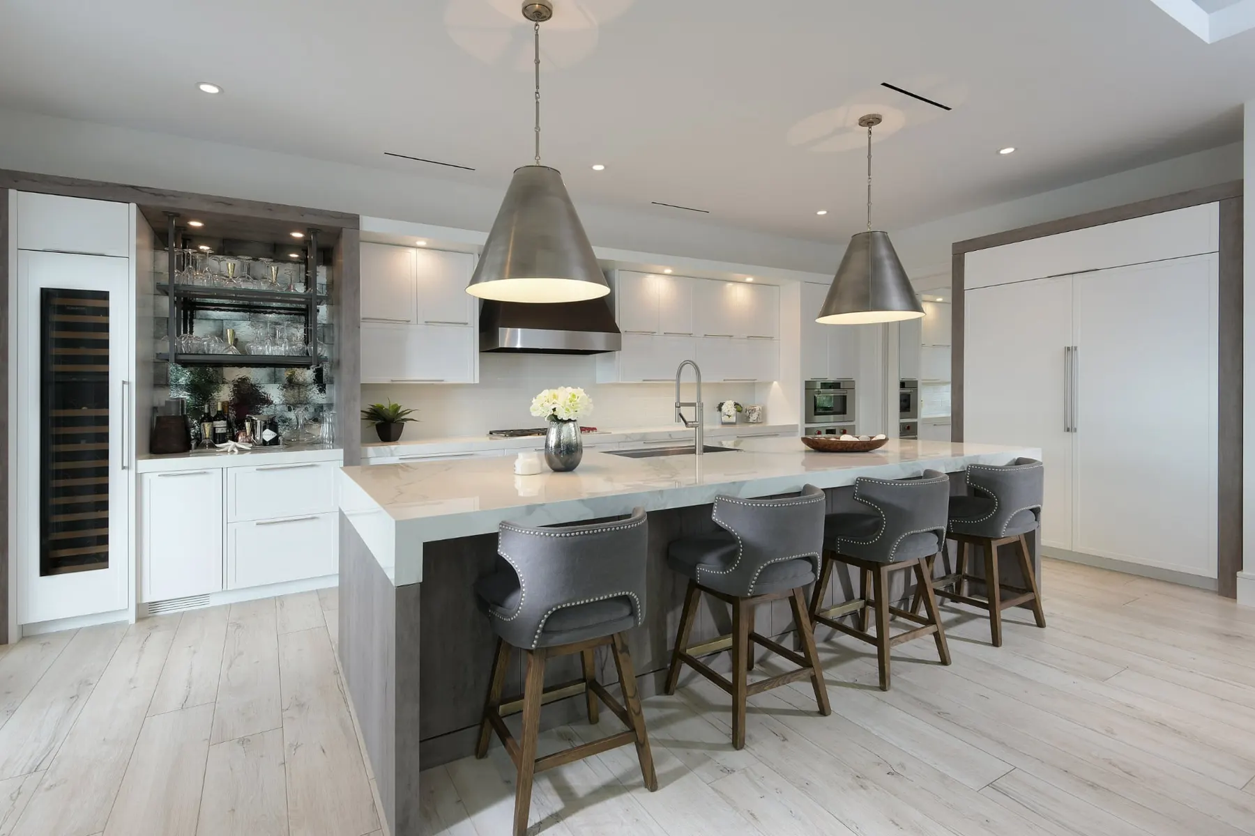 Kitchen with a long island with a stone waterfall edge, four upholstered counter stools, two large metal pendants and a wine column.